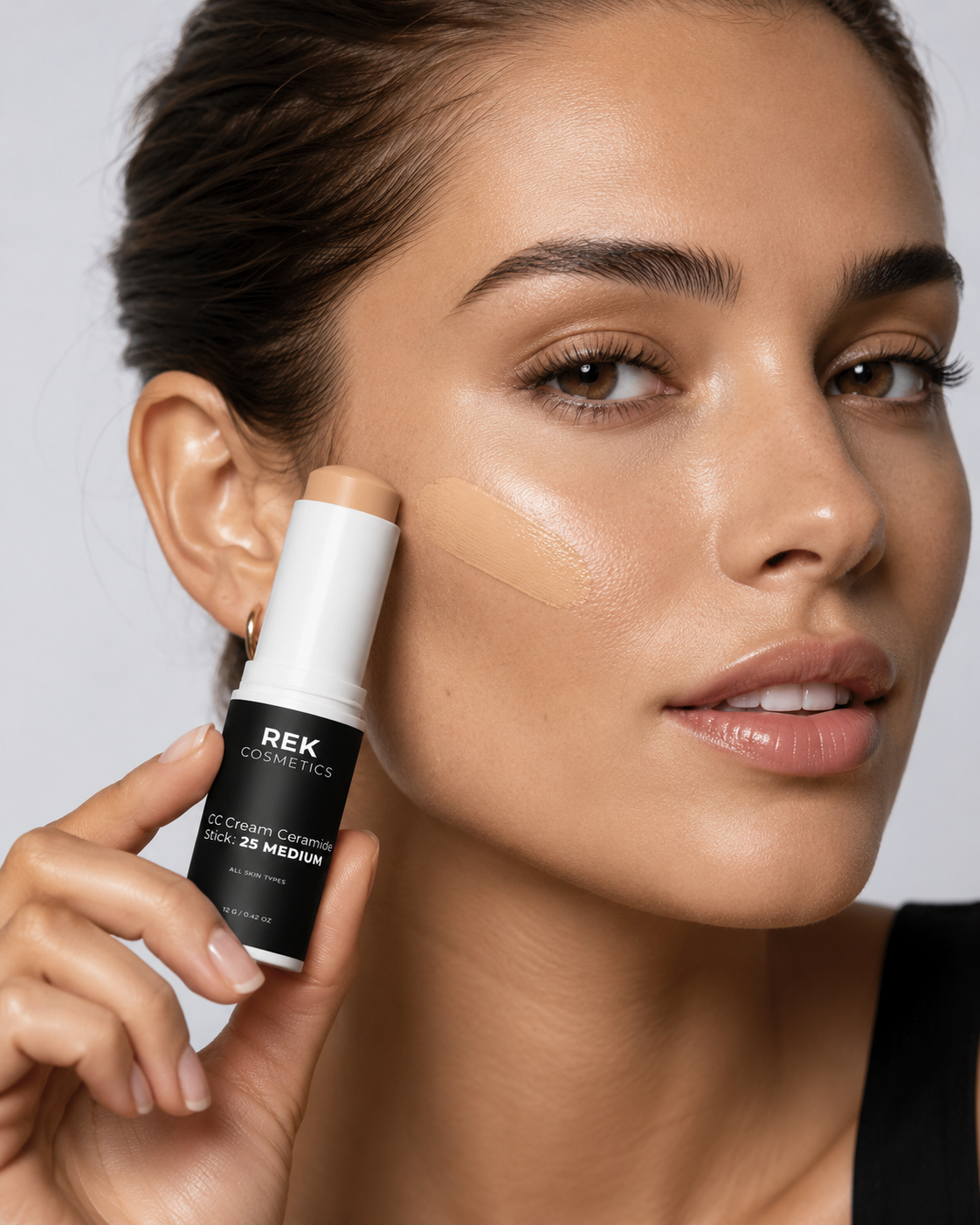 Woman applying REK Cosmetics concealer stick to her face against a neutral background