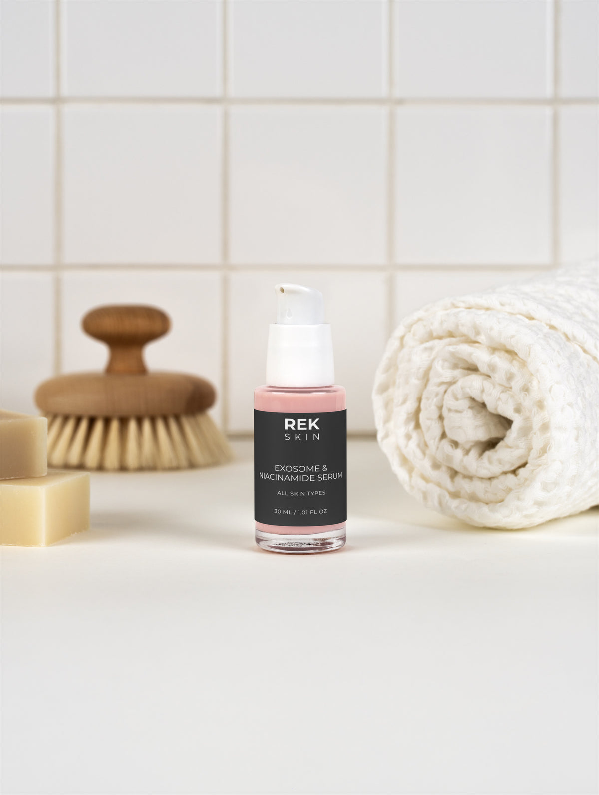 Skincare bottle on a bathroom counter with a brush, soap, and towel in the background.