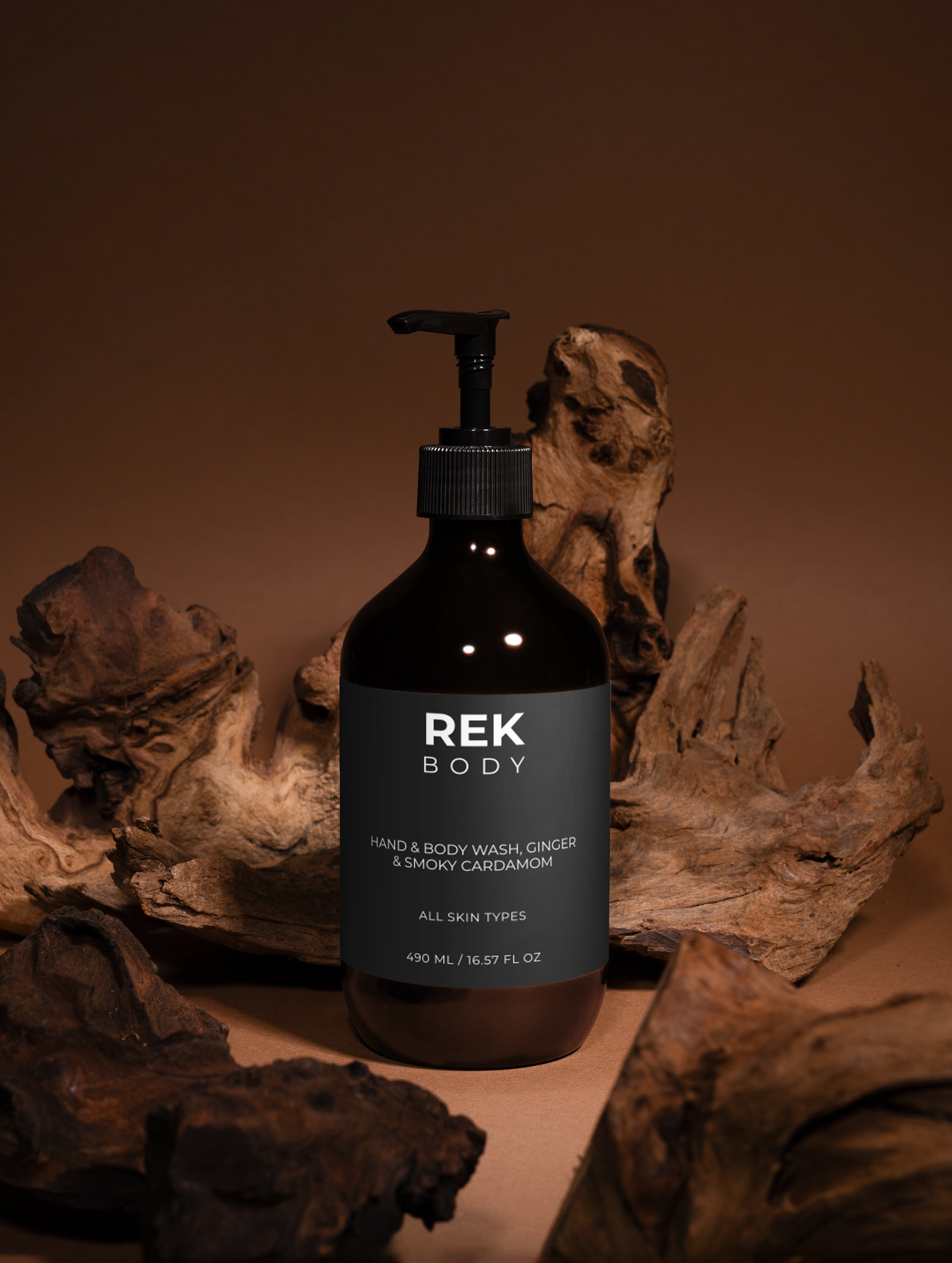 A dark amber pump bottle of REK BODY hand and body wash with a black label, styled on a wooden surface surrounded by natural driftwood against a warm brown background.